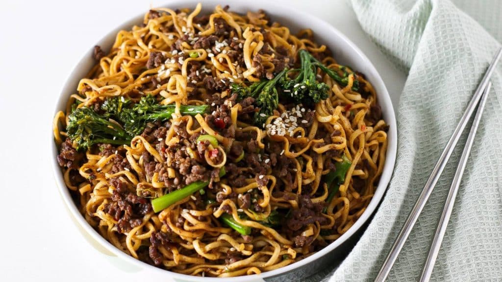 A bowl of ground beef noodle and tenderstem broccoli stir fry with chopsticks and mint green napkin in the background.