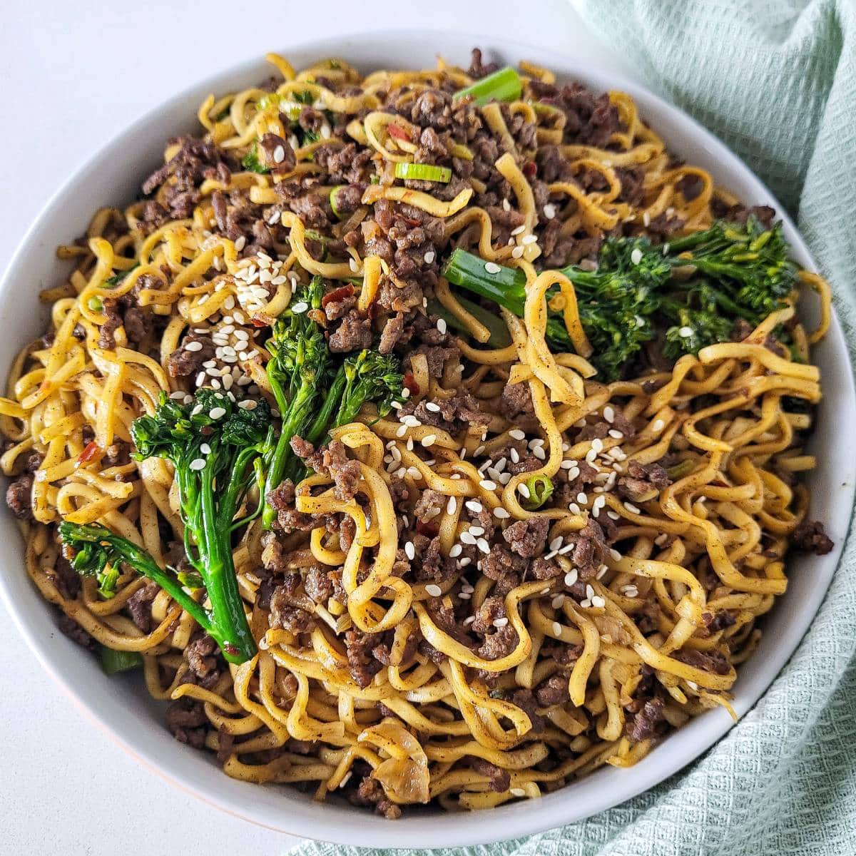 Ground beef noodle stir fry in a white bowl with mint green napkin in the background.