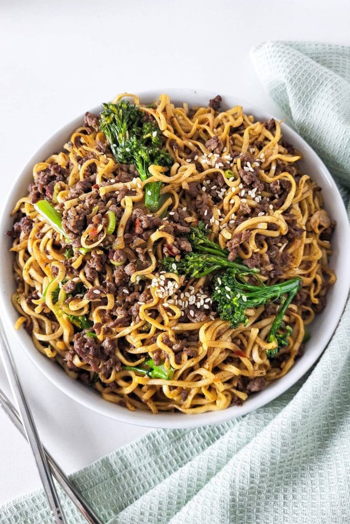A bowl of ground beef noodle stir fry garnished with sesame seeds with chopstick and mint green napkin in the background.