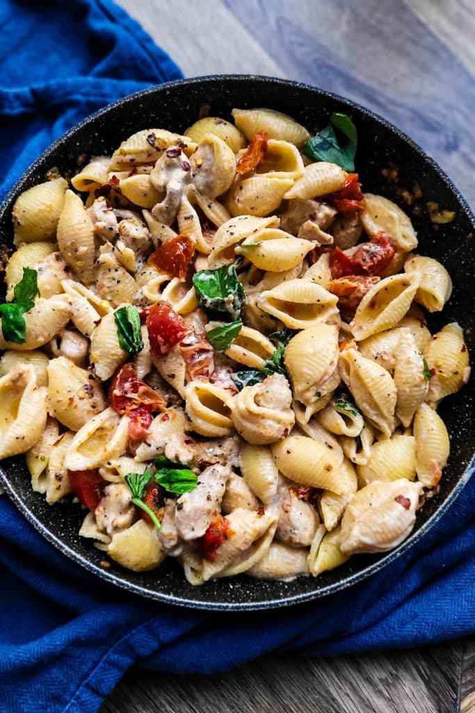 Cooked sun dried tomato pasta with chicken in a skillet with blue napkin in the background.