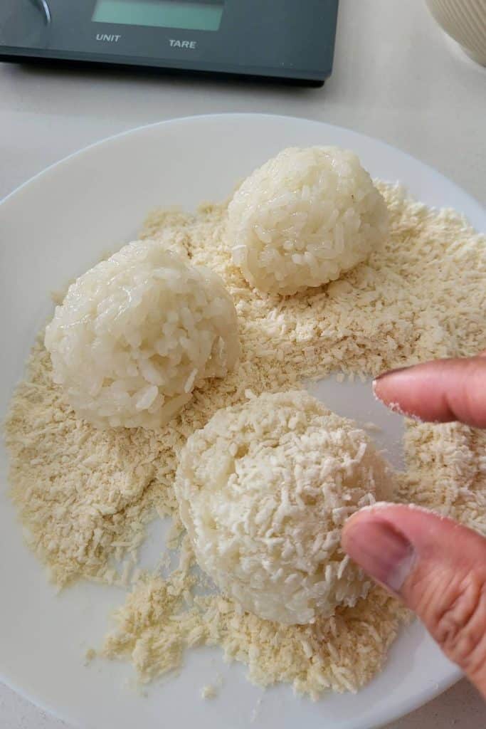 Fingers rolling cooked sweet rice balls over desiccated coconuts.