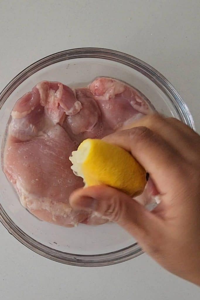 A hand squeezing half a lemon over raw chicken pieces.