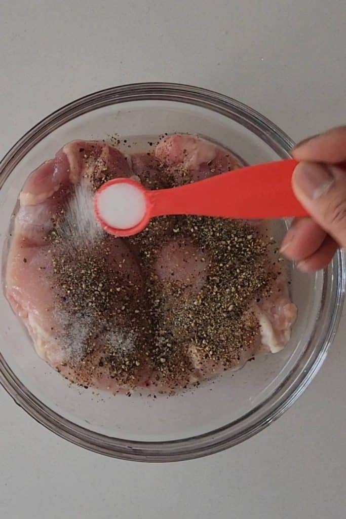 A hand holding a spoon of salt over raw chicken thighs covered with spices.
