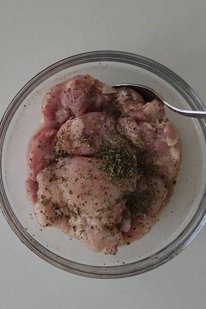 Chicken thighs covered with pepper and dried herbs in a mixing bowl.