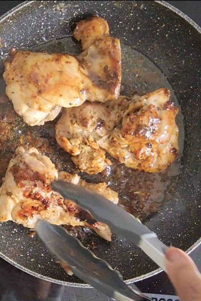 A hand holding a tong over chicken thighs on a skillet.