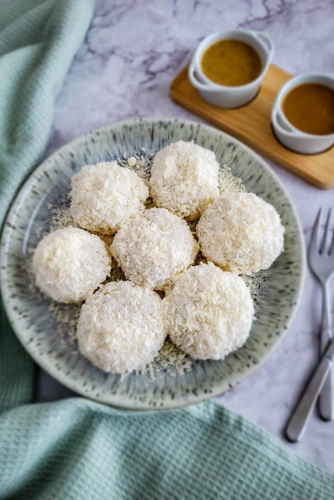 Seven sticky rice balls covered with desiccated coconut on a round plate with two small bowls of peanut sauce, green mint napkin, and forks in the background.