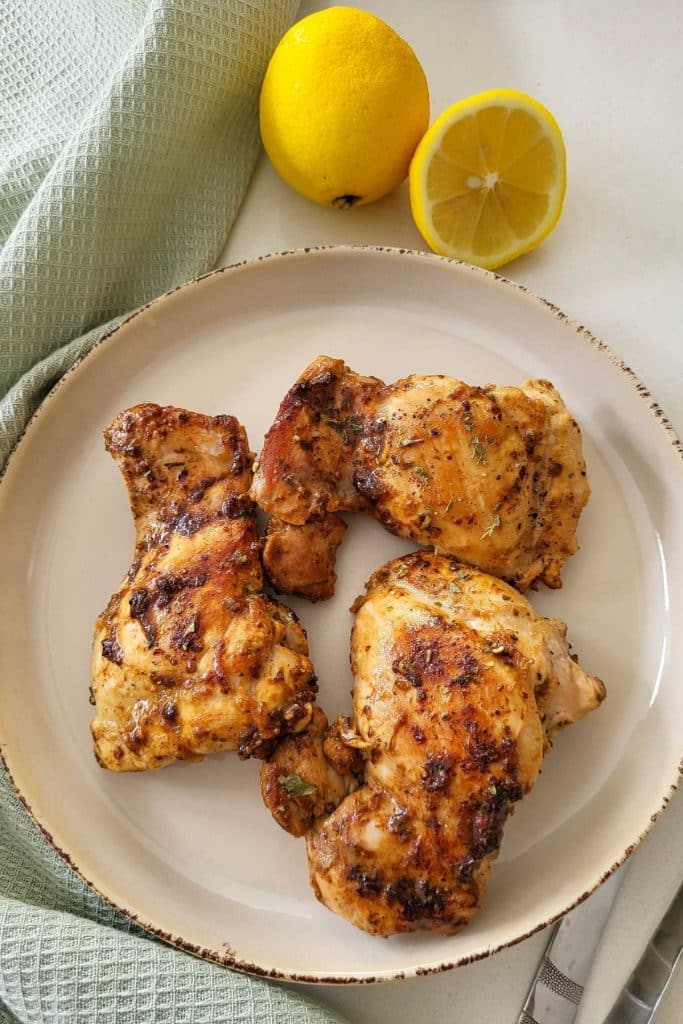 Three pieces of cooked lemon pepper chicken thighs on a plate with lemons and mint green napkin in the background.