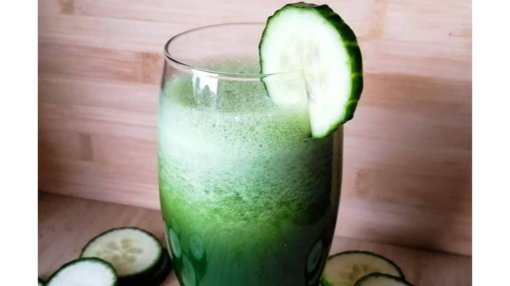 A glass of cucumber and kale smoothie with a slice of cucumber on the glass and a few slices in the background.