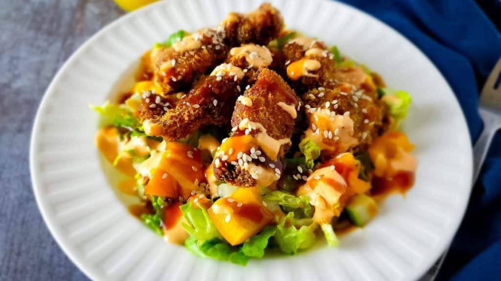 Panko breaded chicken pieces over lettuces, cucumbers, and bell pepper drizzled with katsu sauce and garnished with sesame seeds.