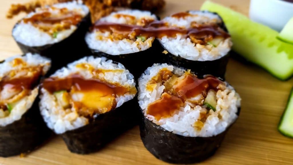 Chicken sushi drizzled with katsu sauce on a wooden chopping board with cucumbers in the background.