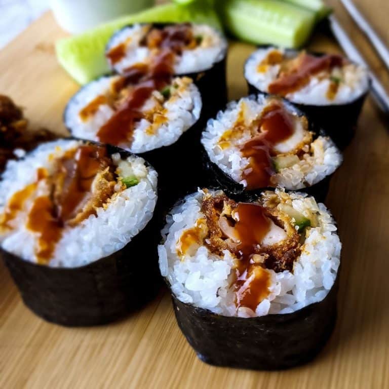 Chicken katsu sushi rolls on a wooden chopping board with cucumbers and chopsticks in the background.