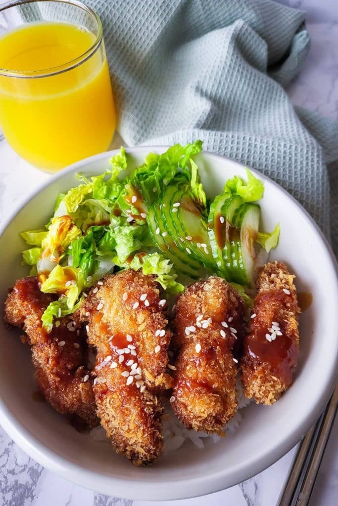 Crispy chicken katsu over seasoned rice with fresh veggies with a glass of orange juice and green mint napkin in the background.