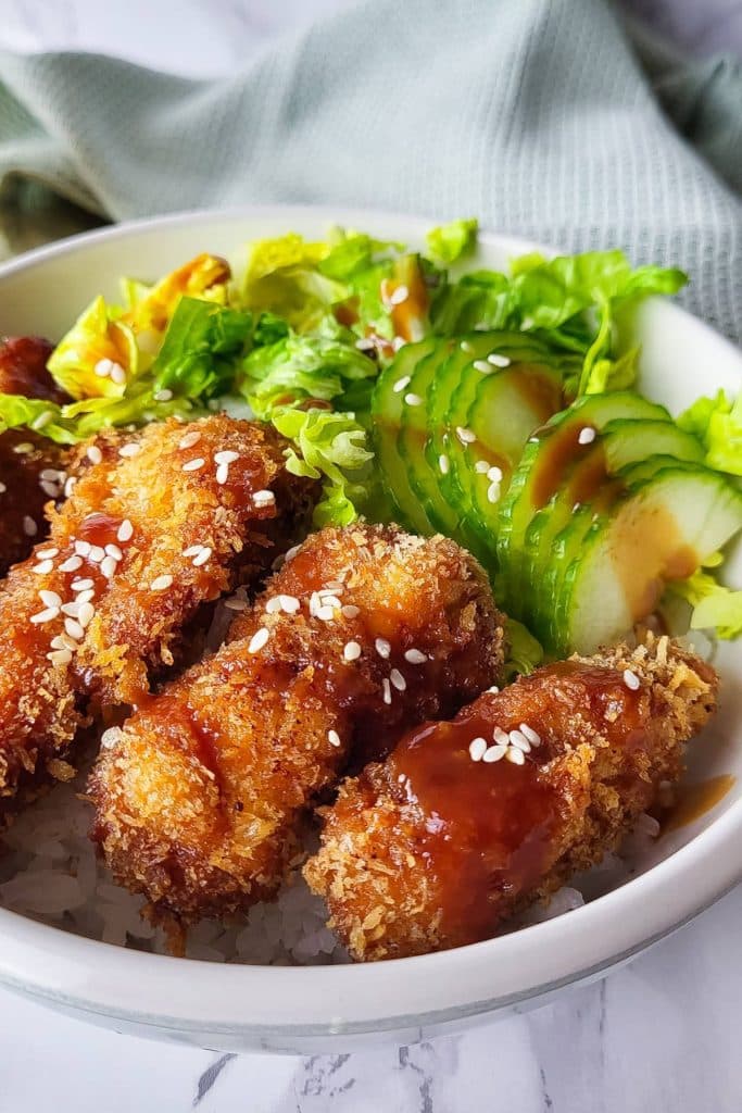 Chicken Katsu pieces in a bowl with seasoned rice and green veggies garnished with sesame seeds.