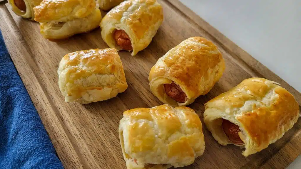 Six puff pastry sausage rolls on a wooden board with blue napkin in the background.