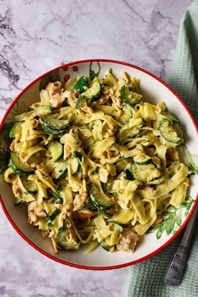 Overhead view of pasta with salmon and zucchini in a wide bowl with green napkin and a fork in the background.