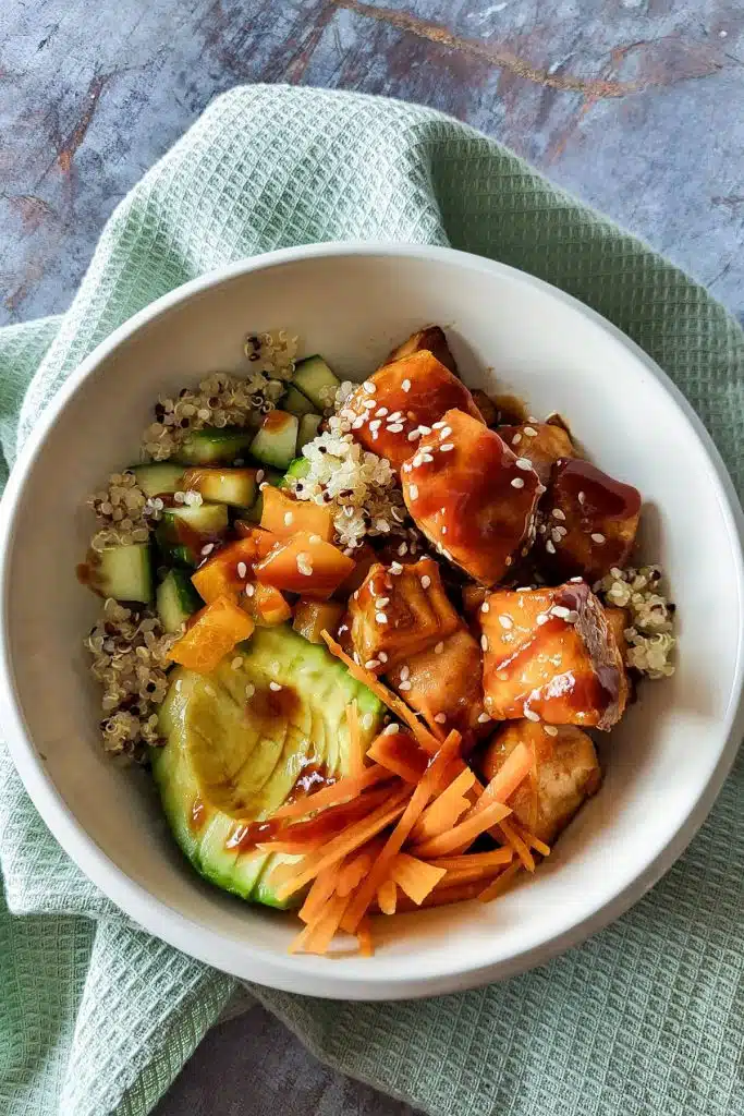 A bowl of salmon in teriyaki sauce with quinoa, avocado, carrots, and cucumbers.