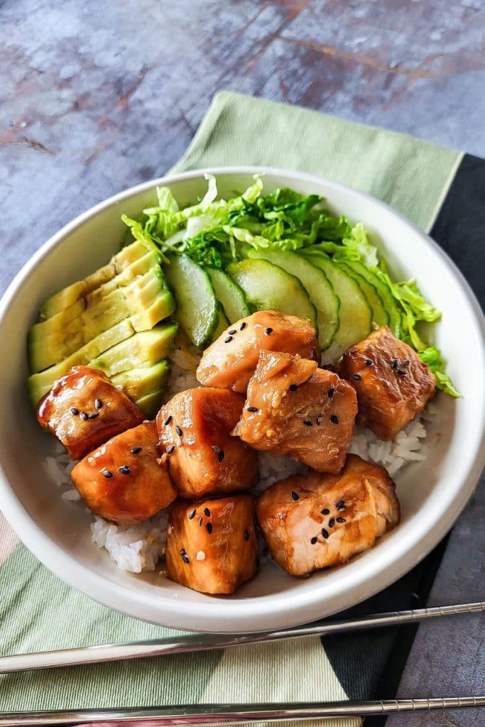 Teriyaki salmon pieces with avocado and cucumber in a bowl with chopsticks in the background.
