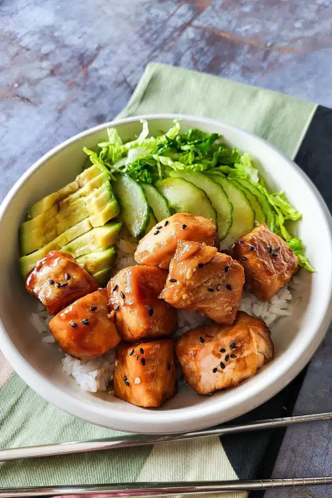 Teriyaki salmon pieces with avocado and cucumber in a bowl with chopsticks in the background.