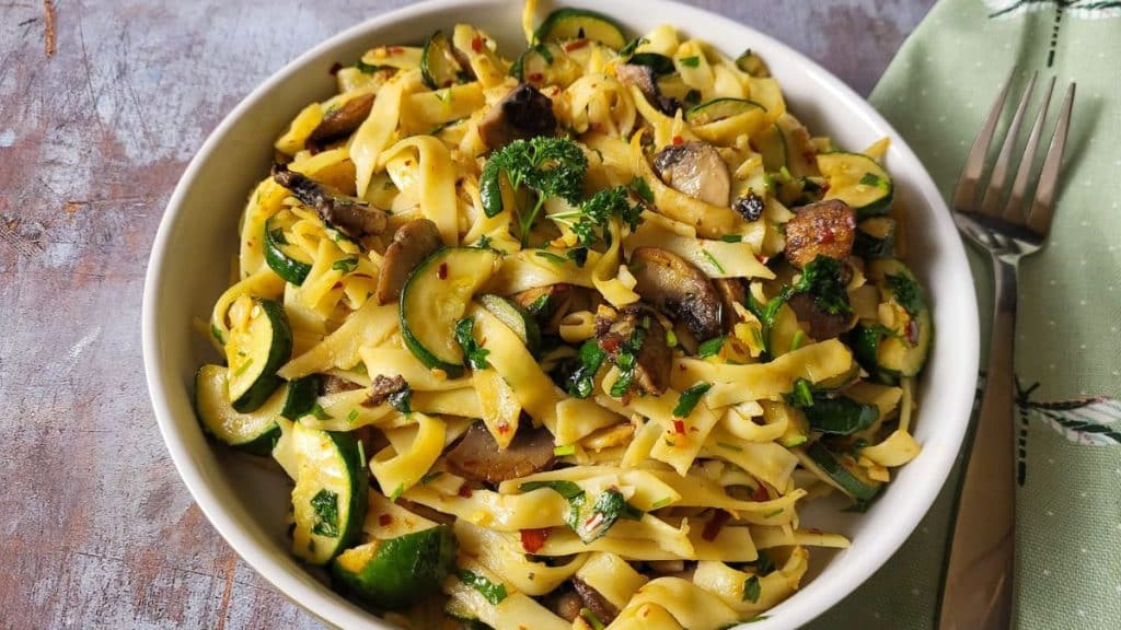 A bowl of zucchini and mushroom pasta with green napkin and a fork in the background.