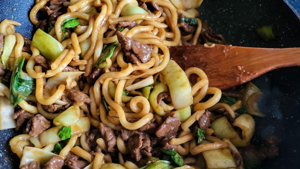 Beef udon noodle stir fry with a wooden slotted turner.